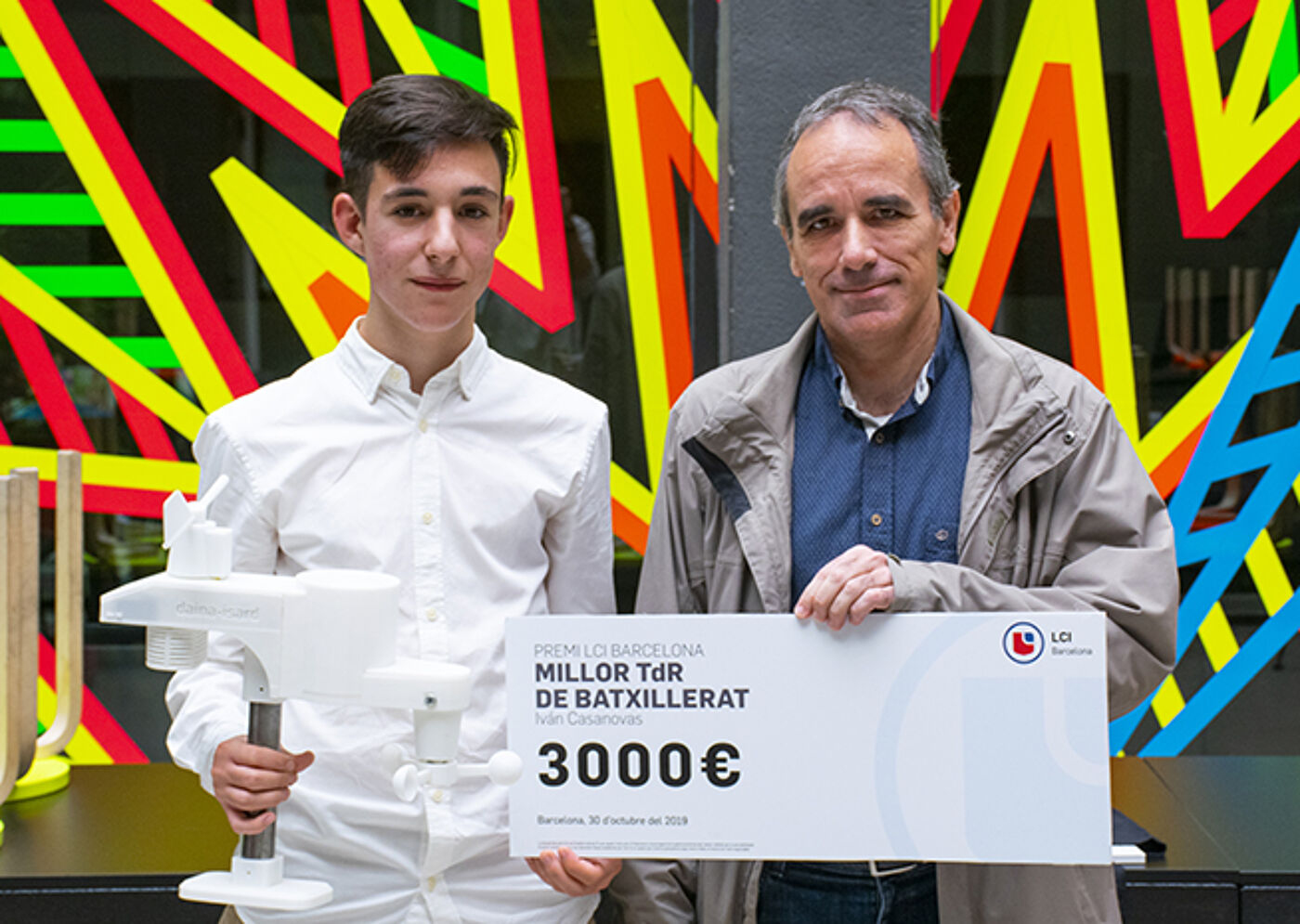 A young individual and an older man pose with a celebratory check for €3000, awarded for the 'Best Baccalaureate Thesis' in Barcelona, dated October 2019.