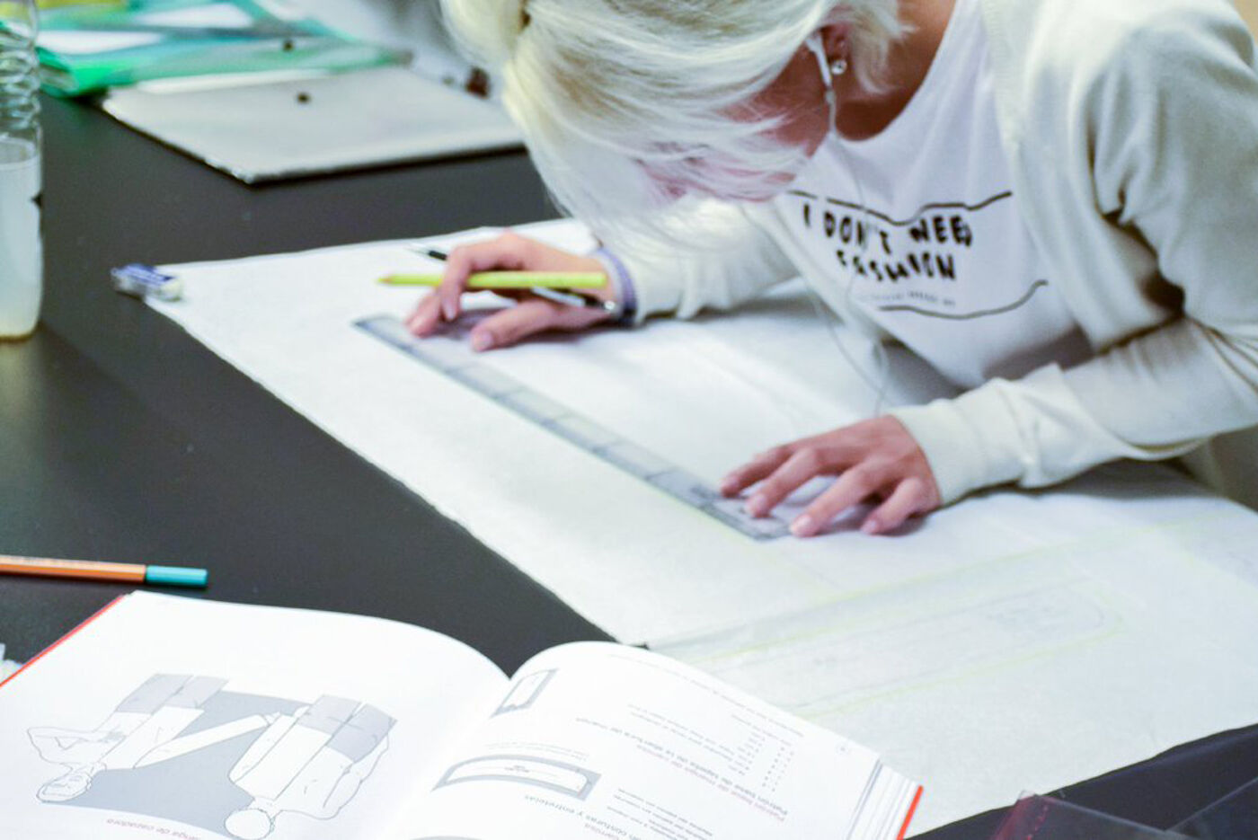 In the image, there is a woman sitting at a table with a book and a ruler. She is working on a project, possibly drawing or studying, and is using the ruler to measure or mark something on the book. The scene suggests that she is engaged in a task that requires precision and attention to detail.