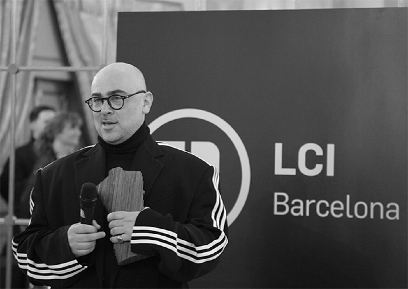 A bald man with glasses, clad in a black turtleneck and jacket with white stripes on the sleeves, holds a microphone at an LCI Barcelona backdrop.