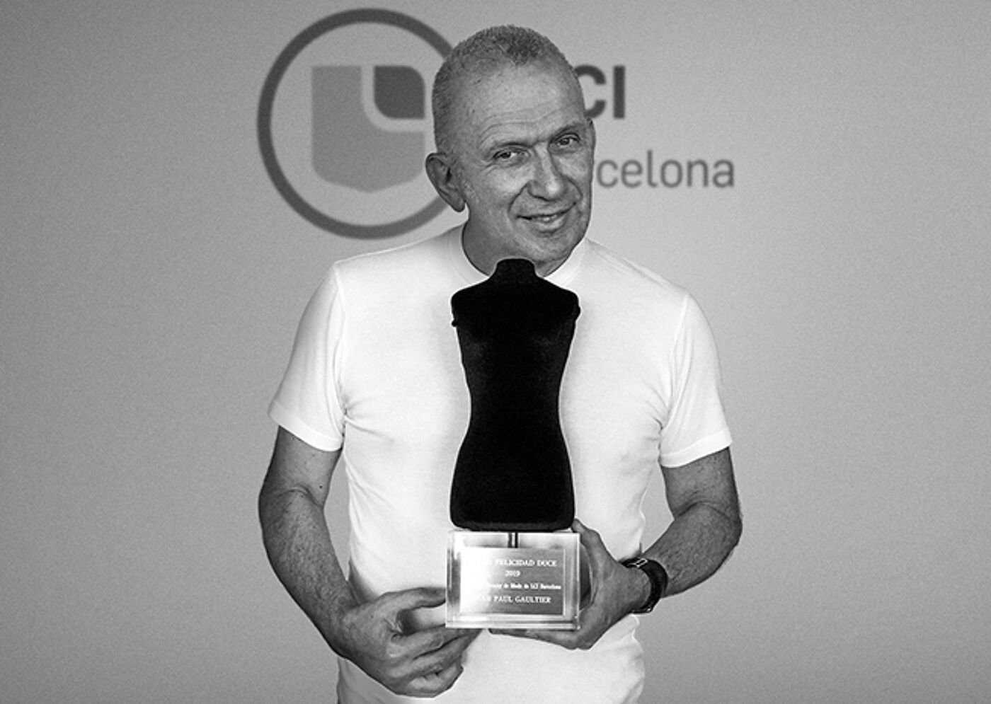 A smiling man in a casual white T-shirt stands in front of a logo, holding a trophy with a plaque that likely bears his name.