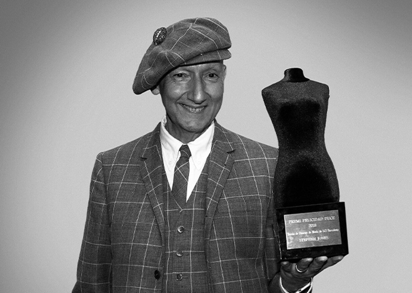 A cheerful man in a plaid suit and beret holds a sculpted award, radiating pride.
