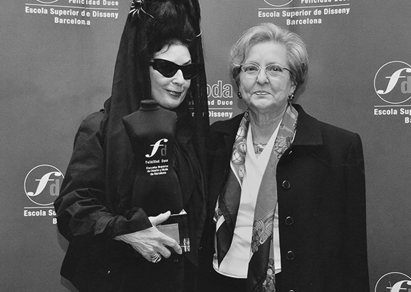 Two women posing at a fashion event; one in avant-garde attire and sunglasses, the other in formal academic dress.