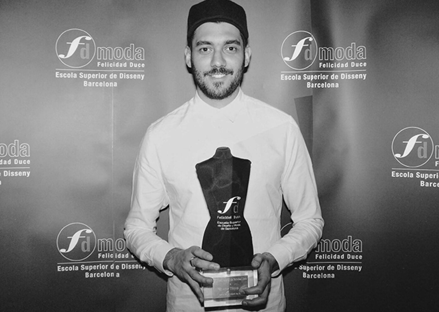 A man in a chef's uniform proudly holds an award at a fashion school event in Barcelona.