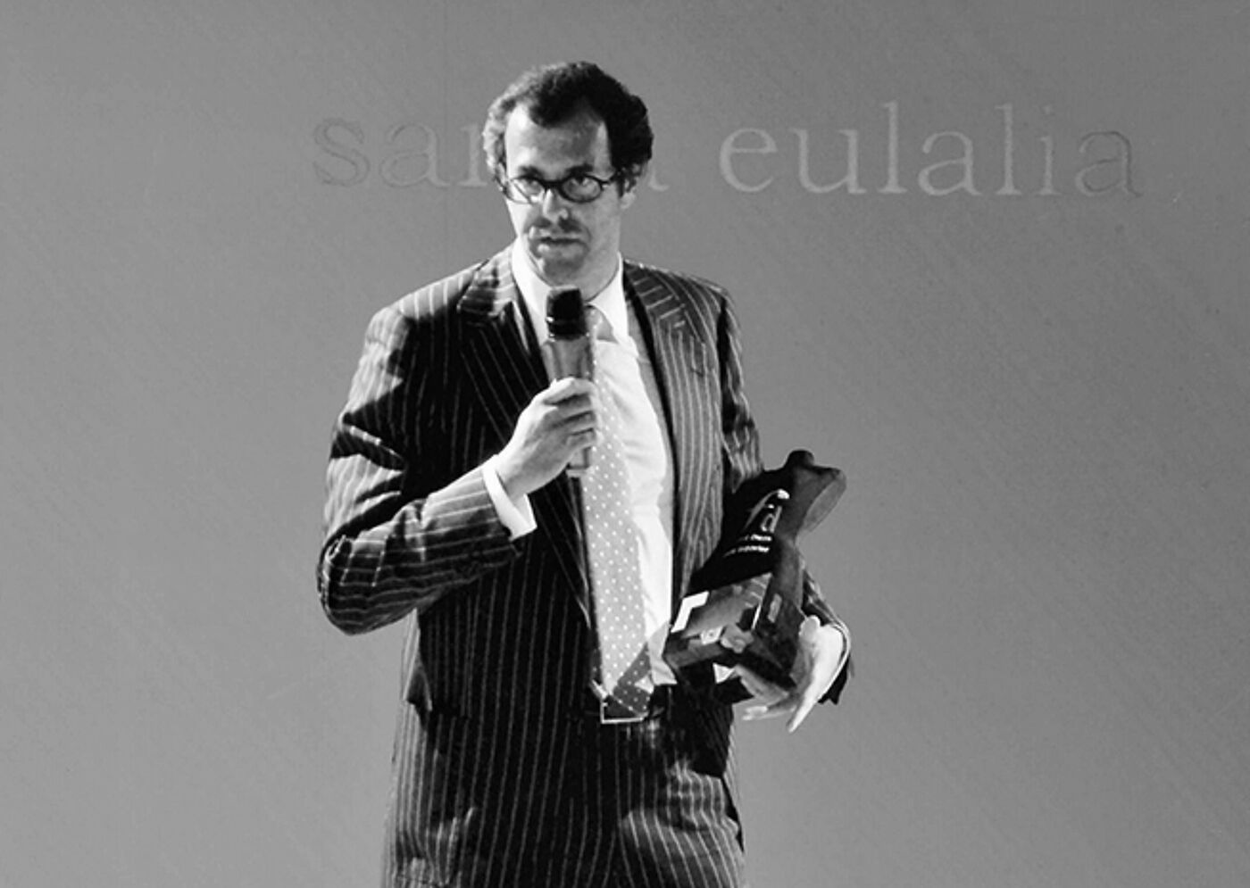 A black and white photo of a man in a striped suit holding a microphone and an award, likely addressing an audience.