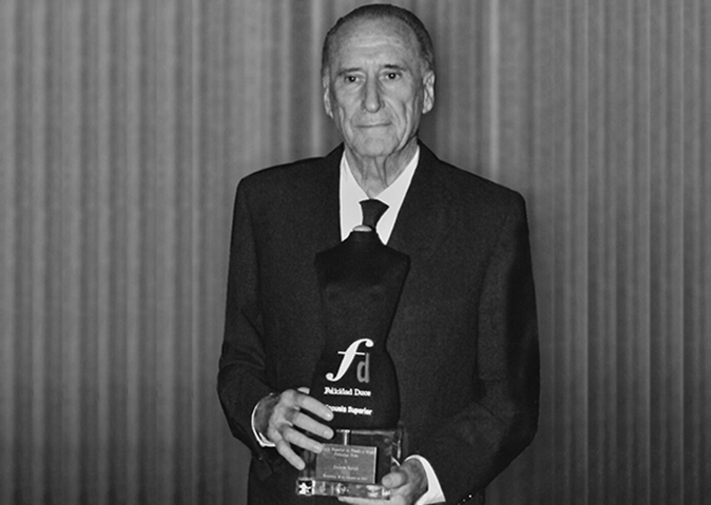 An elderly man in a suit holds a distinguished award, pride evident in his stance.