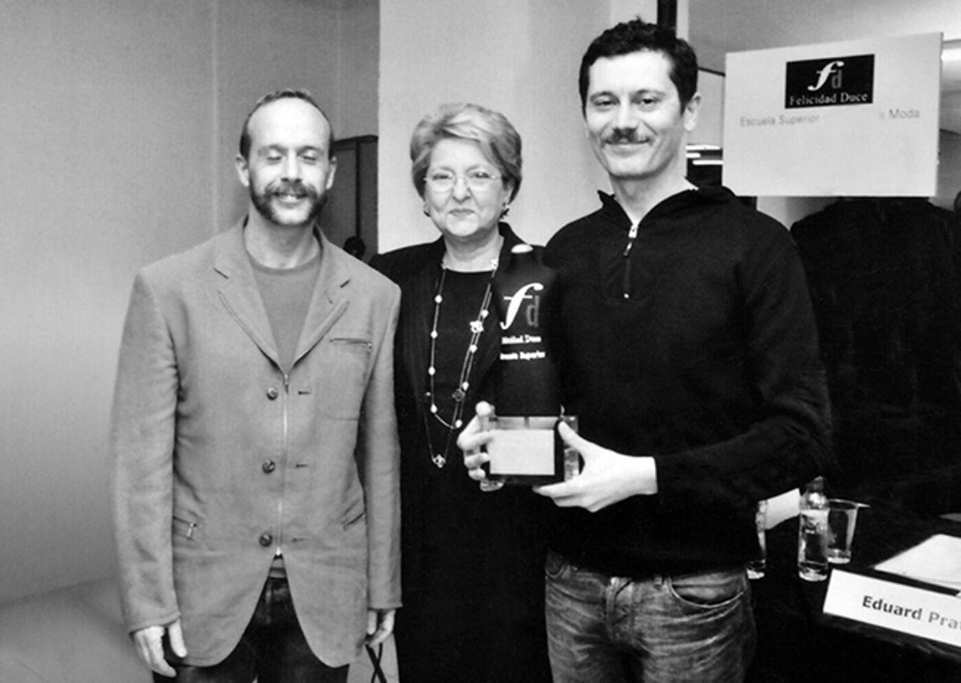 Three individuals posing at a cultural event; a man in the center, possibly an organizer, flanked by two smiling recipients, one holding a small award.