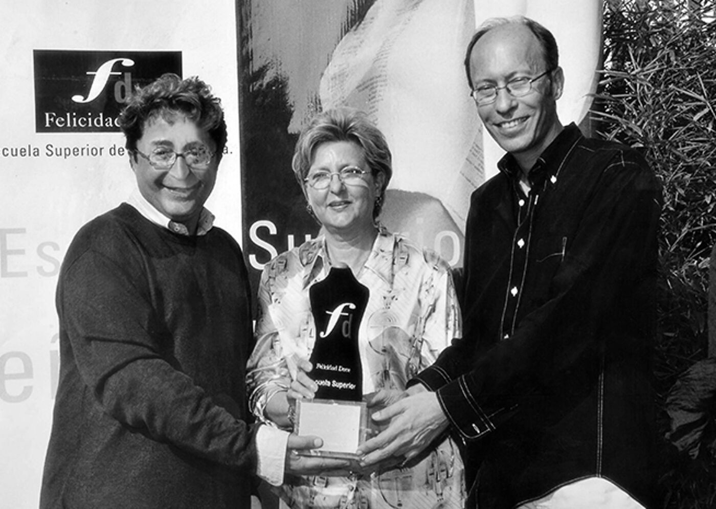 Three individuals pose for a photograph during an award presentation; a woman in the center holds a trophy with an inscription, flanked by two smiling men.