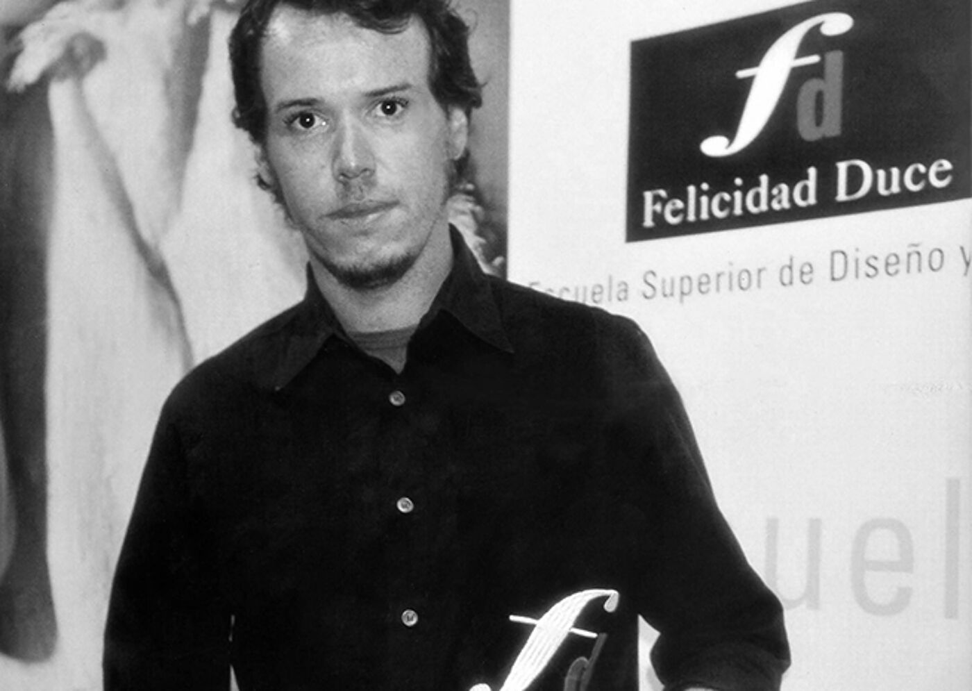 A young man in a black shirt poses at a fashion design school event, a sign "Felicidad Duce" is visible in the background.