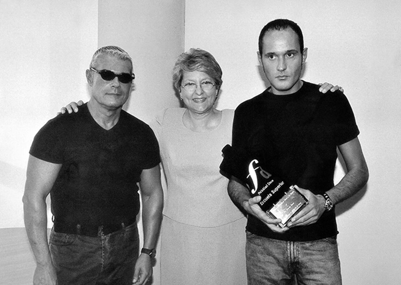 An elderly couple and a younger man pose together, the latter holding a clear award.