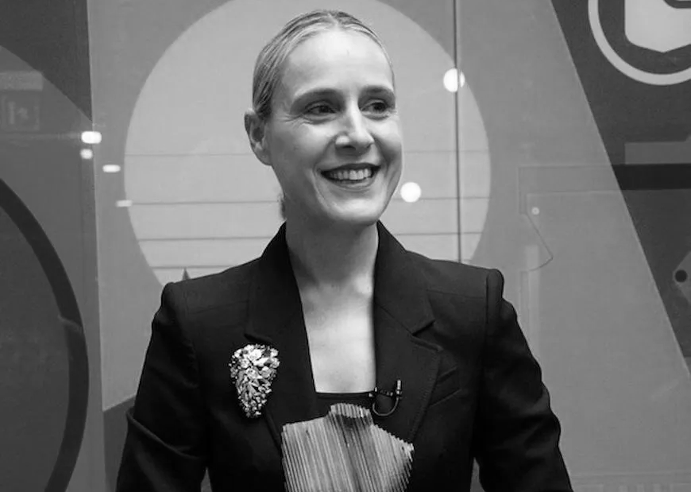 Black-and-white image of a woman with light hair tied back, smiling while wearing a formal blazer. She has a decorative brooch on her left lapel and a microphone clipped to her outfit. The abstract background features geometric shapes and soft lighting.