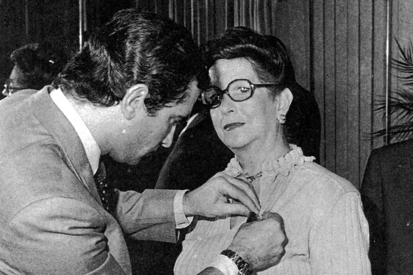 In the picture, we have a man and a woman standing next to each other. The man is fixing a pin or a brooch on the woman's clothing, possibly her neck or her dress. The woman is wearing glasses, and the man is adjusting the pin to ensure it is properly placed and secure.