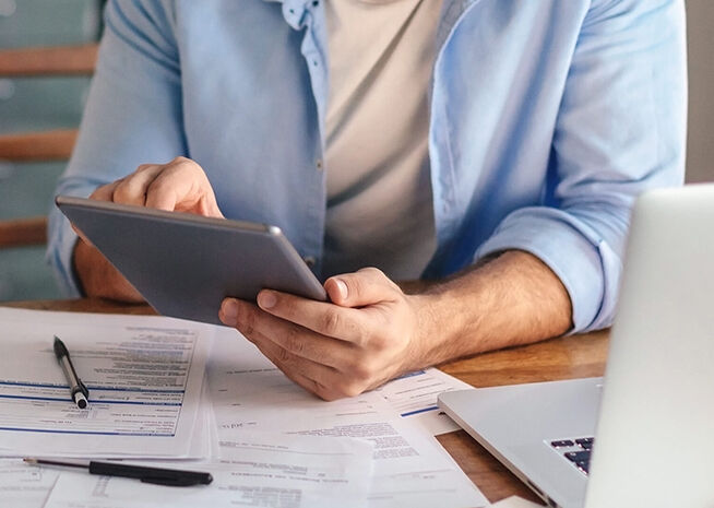 A man in a light blue shirt attentively examines a tablet amidst papers and a laptop on a desk.