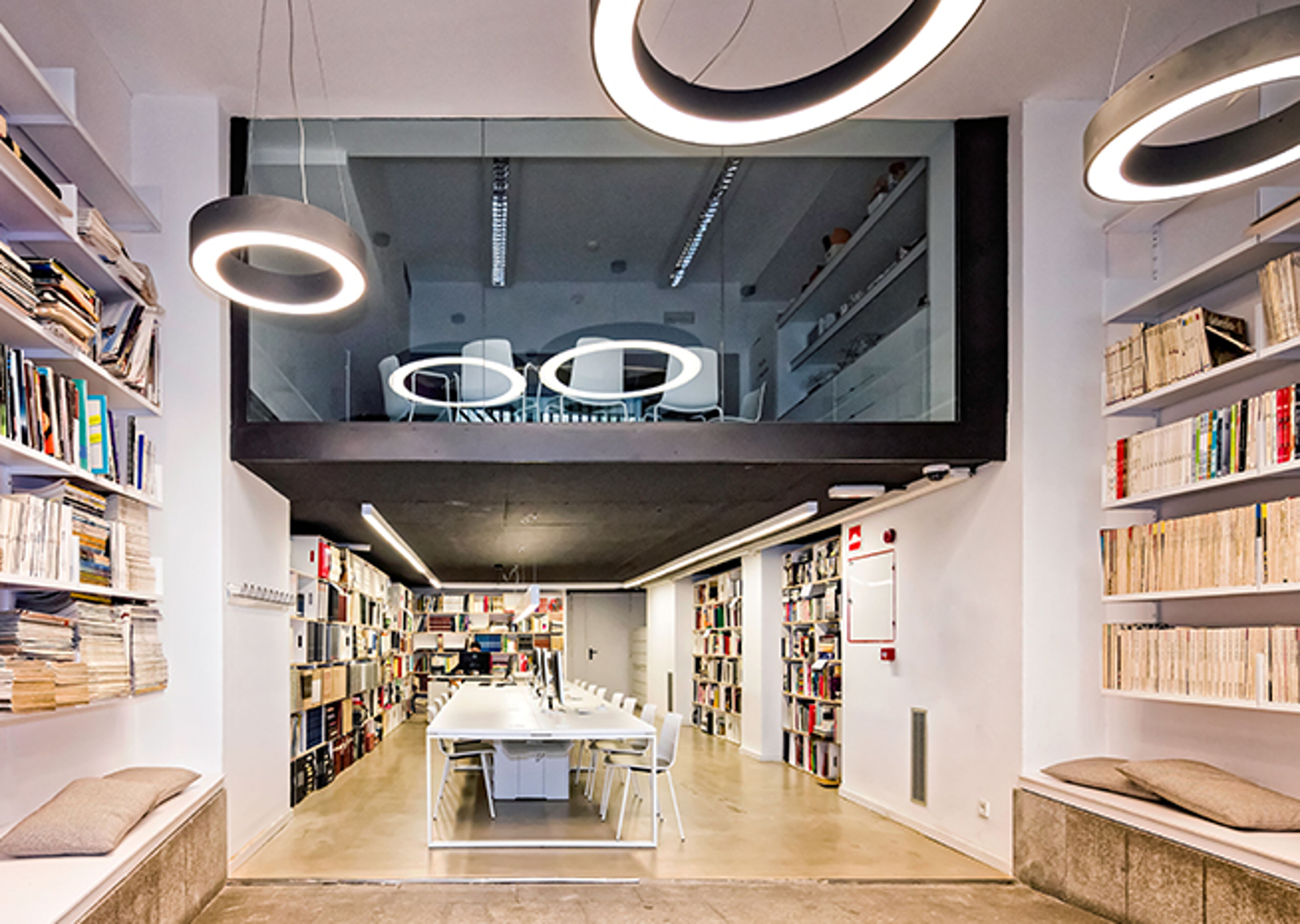 A contemporary library space with circular lighting fixtures, a central communal table, and wall-to-wall bookshelves.