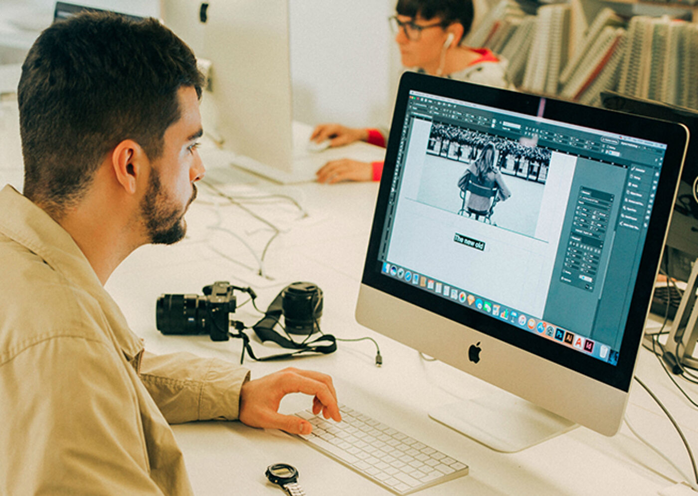 A graphic designer works on a photo editing project on a desktop computer, with camera equipment nearby.