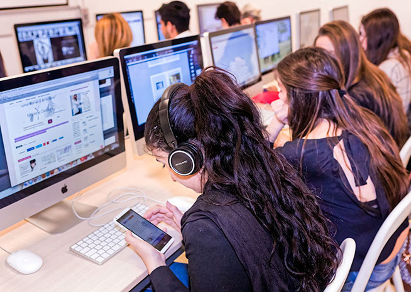 Students focused on their work in a computer lab, using desktops and personal devices with headphones.