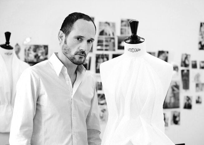 A focused fashion designer stands beside a mannequin in a studio, with design sketches in the background.