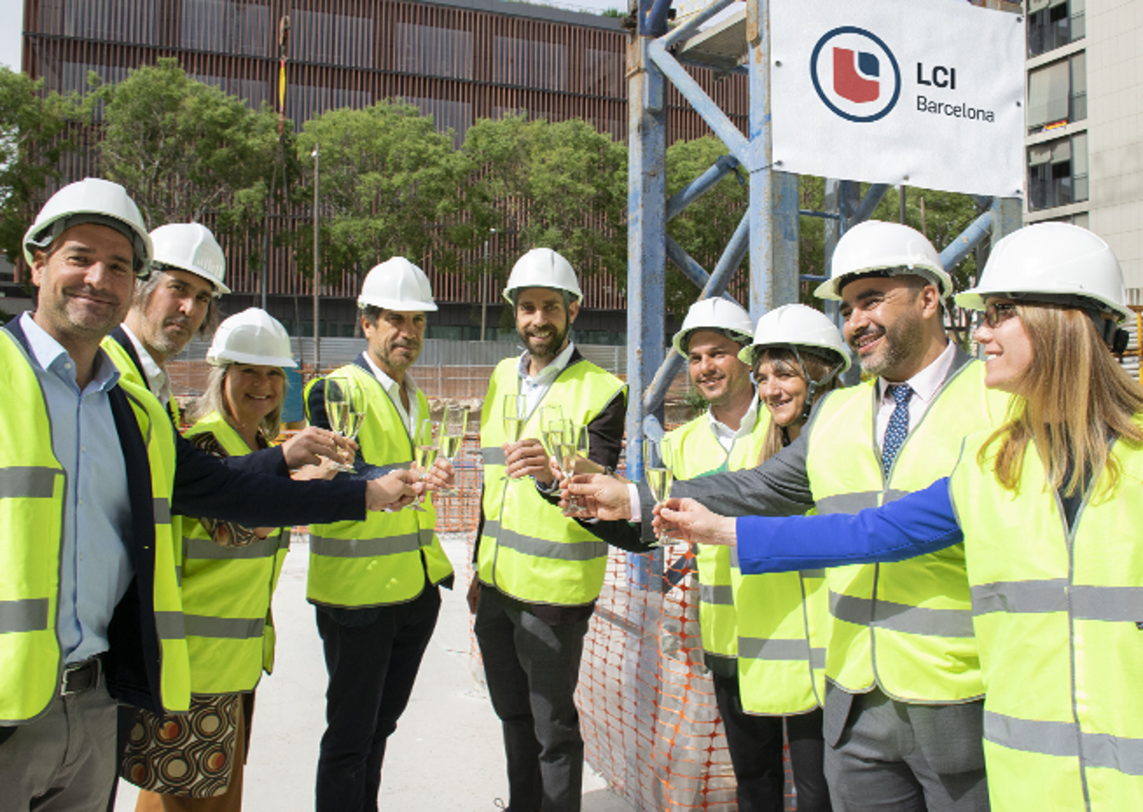  Grupo de profesionales con cascos y chalecos reflectantes celebrando con un brindis al aire libre en un sitio de construcción, cartel de LCI Barcelona al fondo.