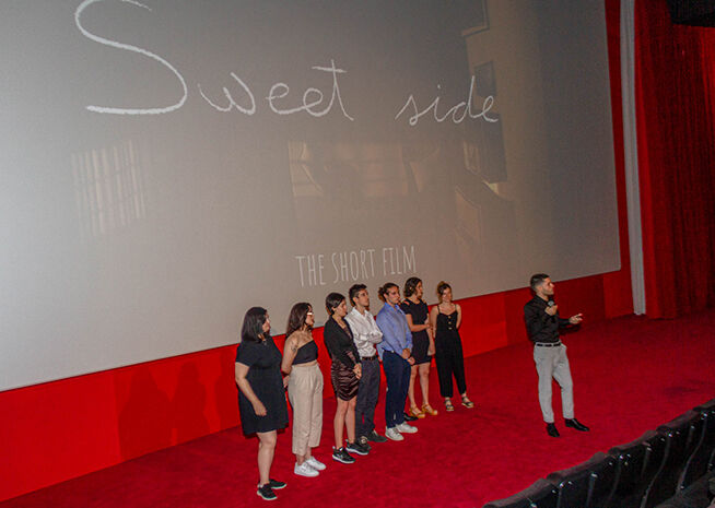 Un grupo está en el escenario de un cine durante el estreno de 'Sweet Side: El Cortometraje', interactuando con la audiencia.