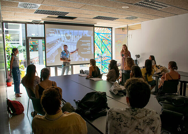 A presenter gesturing towards a screen in a brightly lit classroom with attentive students.