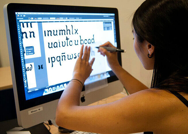 Un(a) diseñador(a) gráfico(a) usa un lápiz óptico en una tableta de dibujo digital conectada a una computadora de escritorio, editando elementos tipográficos.
