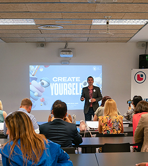 A speaker presents to a seated audience in a conference room with a "CREATE YOURSELF" slide on screen.
