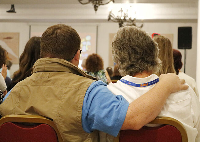 Two people seated, one with an arm around the other, at a conference hall with audience clapping in the background.