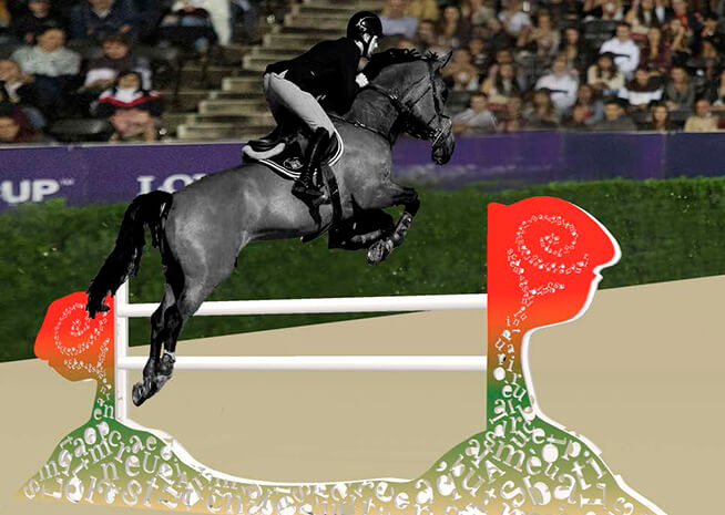 A gray horse and rider in mid-jump over a white showjumping obstacle against a blurred audience background.
