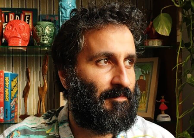 A man with a thick beard and contemplative expression, surrounded by colorful decor and books.