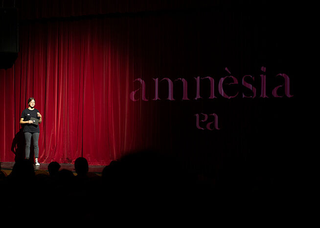 Un intèrpret a l'escenari sota els focus amb la paraula 'amnèsia' projectada al fons, creant una atmosfera de drama i expectació.