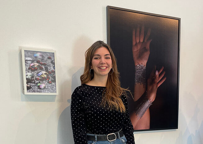Una jove somrient amb una samarreta de punts està al costat d'una fotografia de mans amb joies, afegint un toc personal a la mostra d'art.