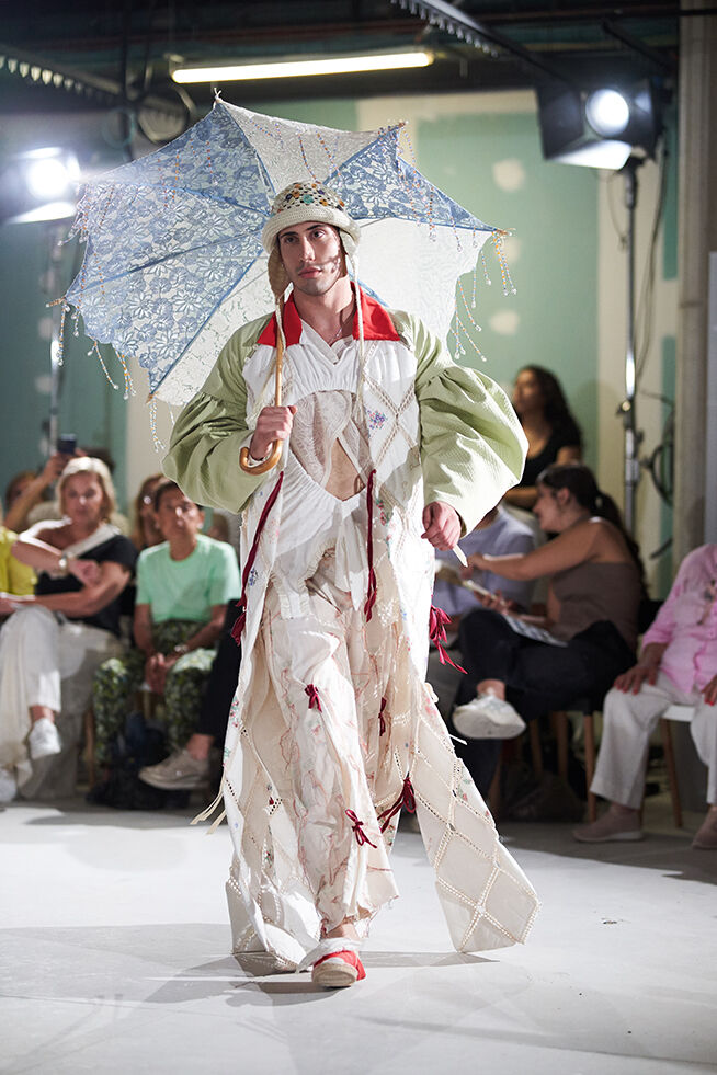Modelo en pasarela exhibiendo un atuendo audaz y ecléctico con un gran paraguas estampado, prendas superpuestas y detalles de cinta, emitiendo una vibra creativa y vanguardista.