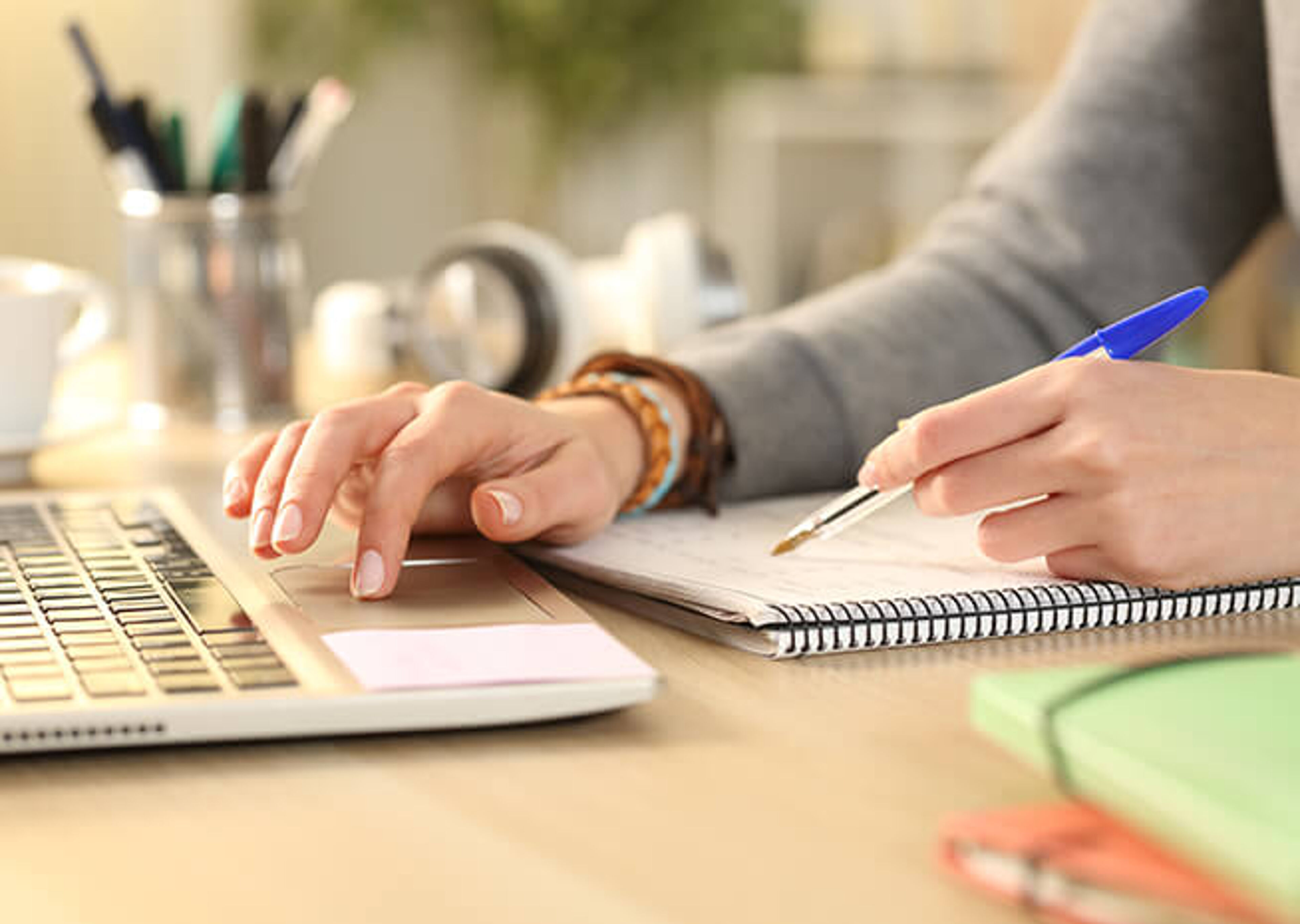 Primer plano de las manos de una persona trabajando, escribiendo en una libreta junto a un portátil en un escritorio bien organizado.