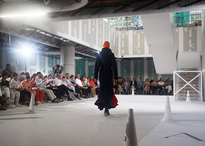 A model walking away from the audience on a fashion show runway, wearing a long black garment with a dramatic red hem.