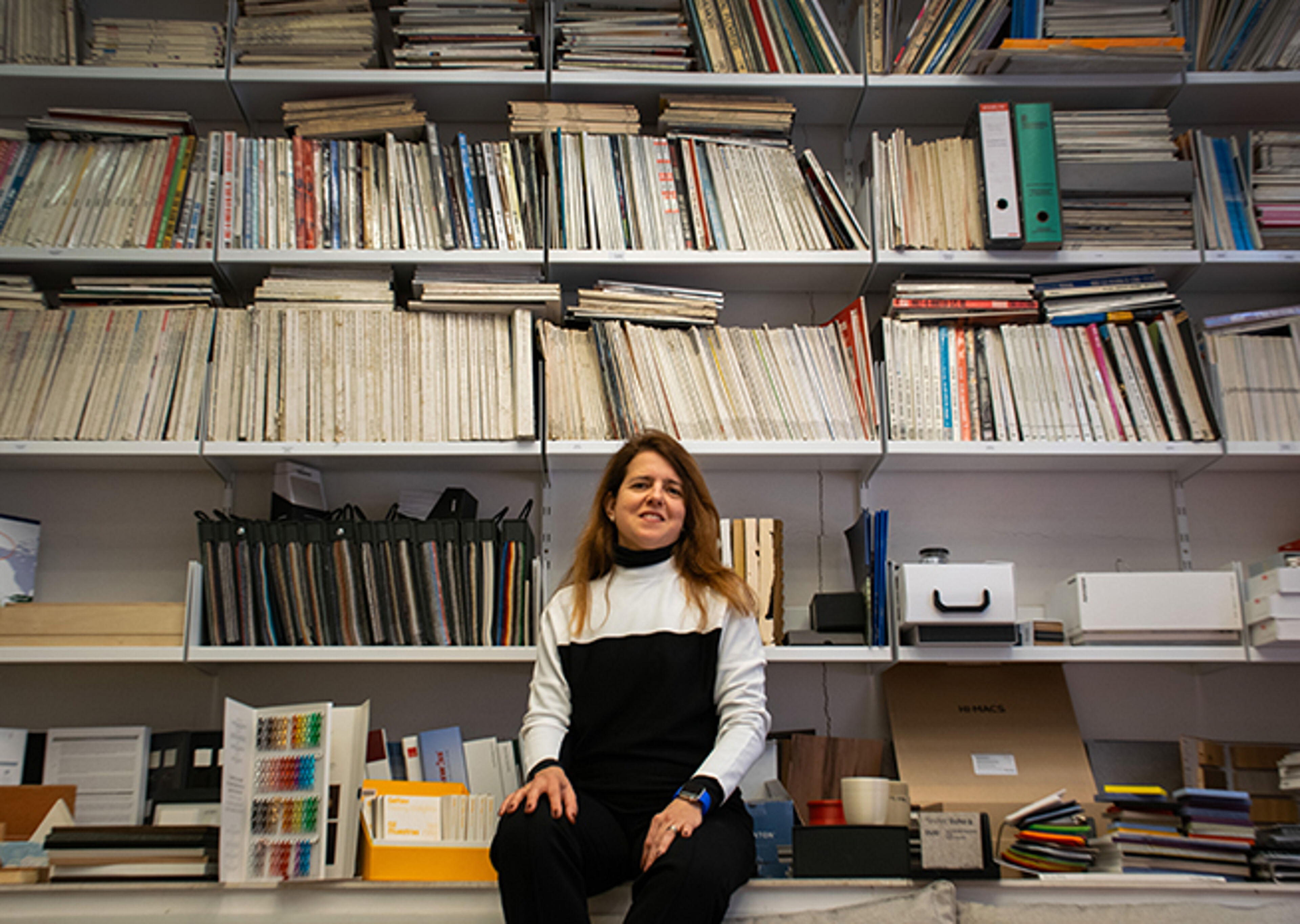 A person sitting in front of a well-stocked bookshelf filled with files and publications in an academic or professional setting.