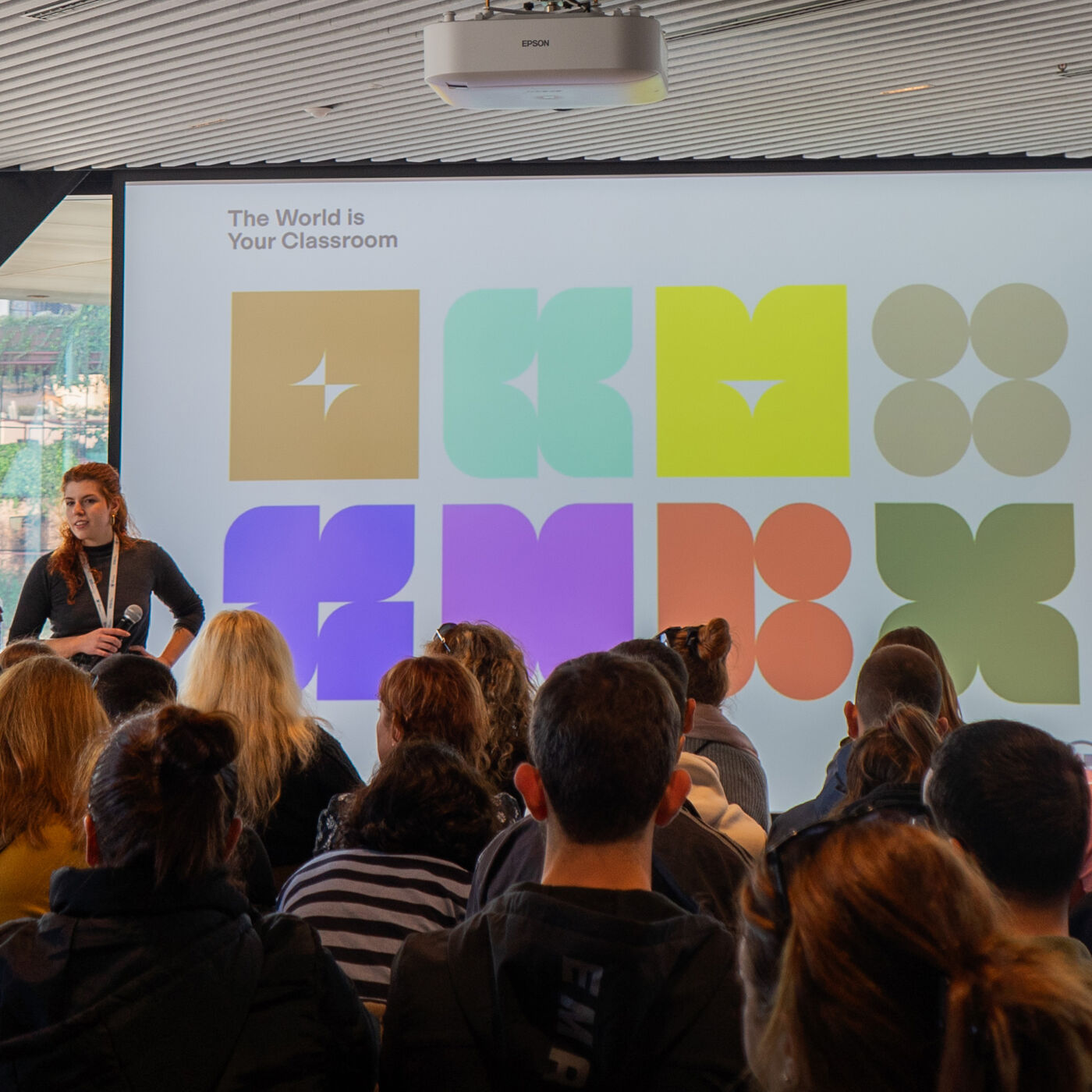 A diverse group of people are seated in an auditorium facing a screen displaying a colorful logo and the words "The World is Your Classroom".