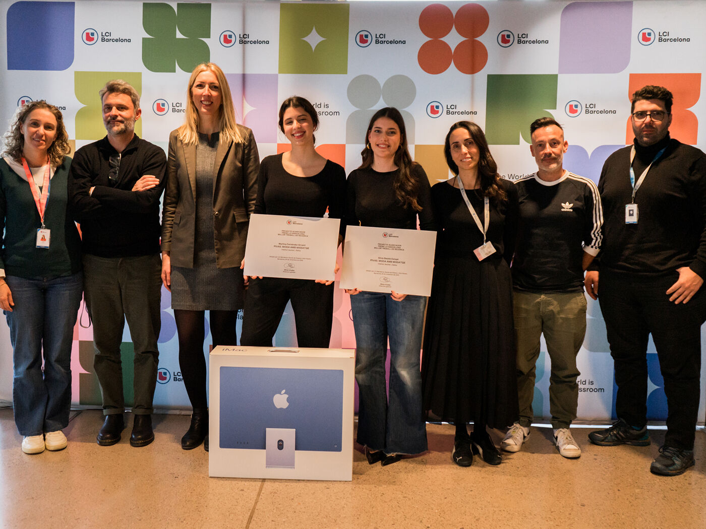 A group of nine people are posing for a photo in front of a colorful backdrop. Several of them are holding certificates, and there's an Apple computer on a stand in front of them.