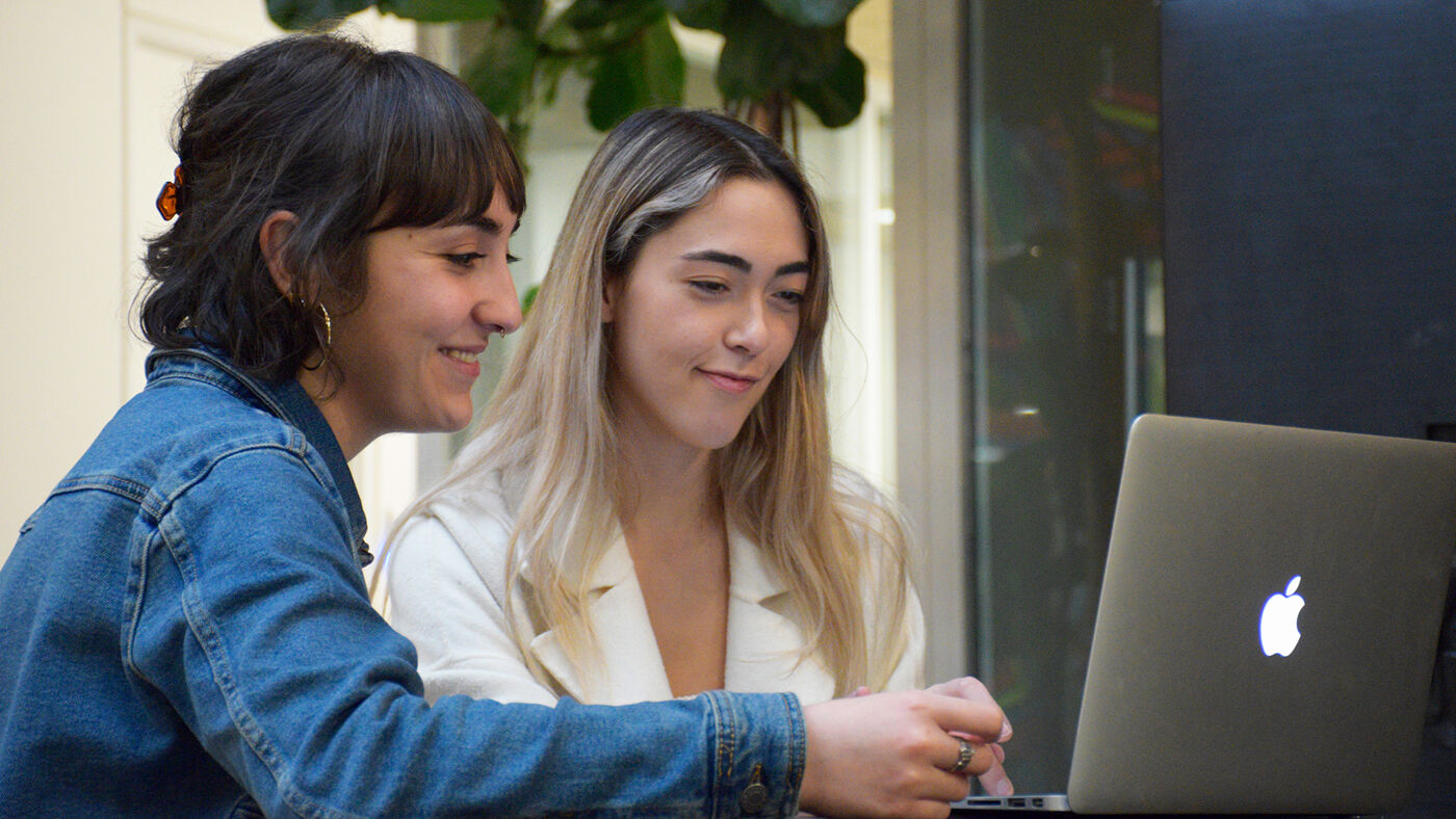 Two women are looking at a laptop screen. One woman is pointing at the screen, while the other is smiling. They appear to be collaborating or working together on something.