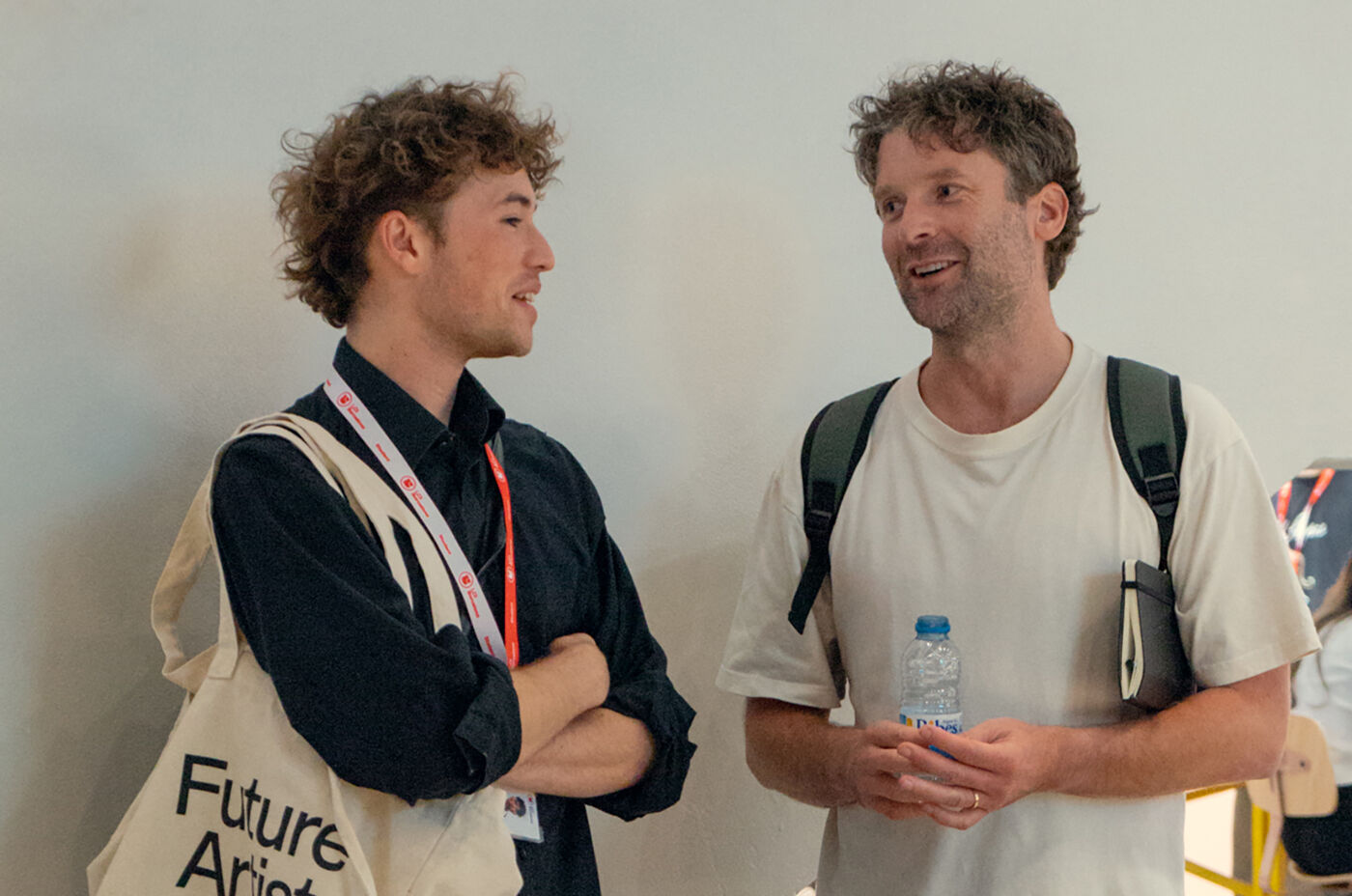 Two men are engaged in a conversation. The man on the left is wearing a black shirt and has a tote bag over his shoulder. The man on the right is wearing a white t-shirt and has a backpack.