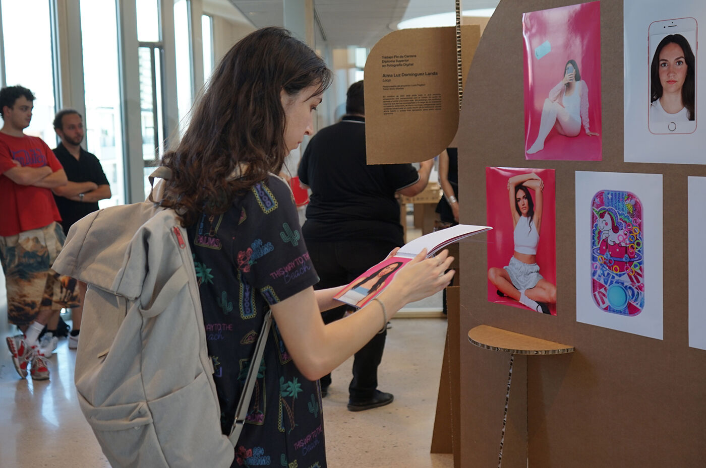A woman with a backpack is looking at artwork displayed on a wall. There are other people in the background, and the artwork includes photographs and illustrations.