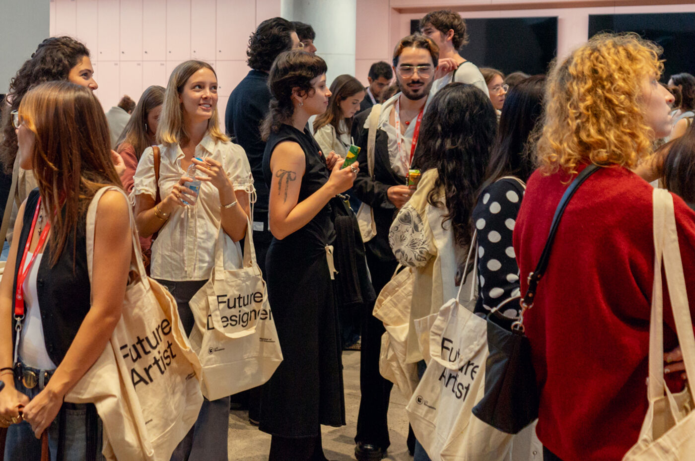 A group of people are gathered in what appears to be an art gallery or event space. Several individuals are holding tote bags with the words "Future Artist" or "Future Designer" printed on them. The atmosphere seems lively and engaged.