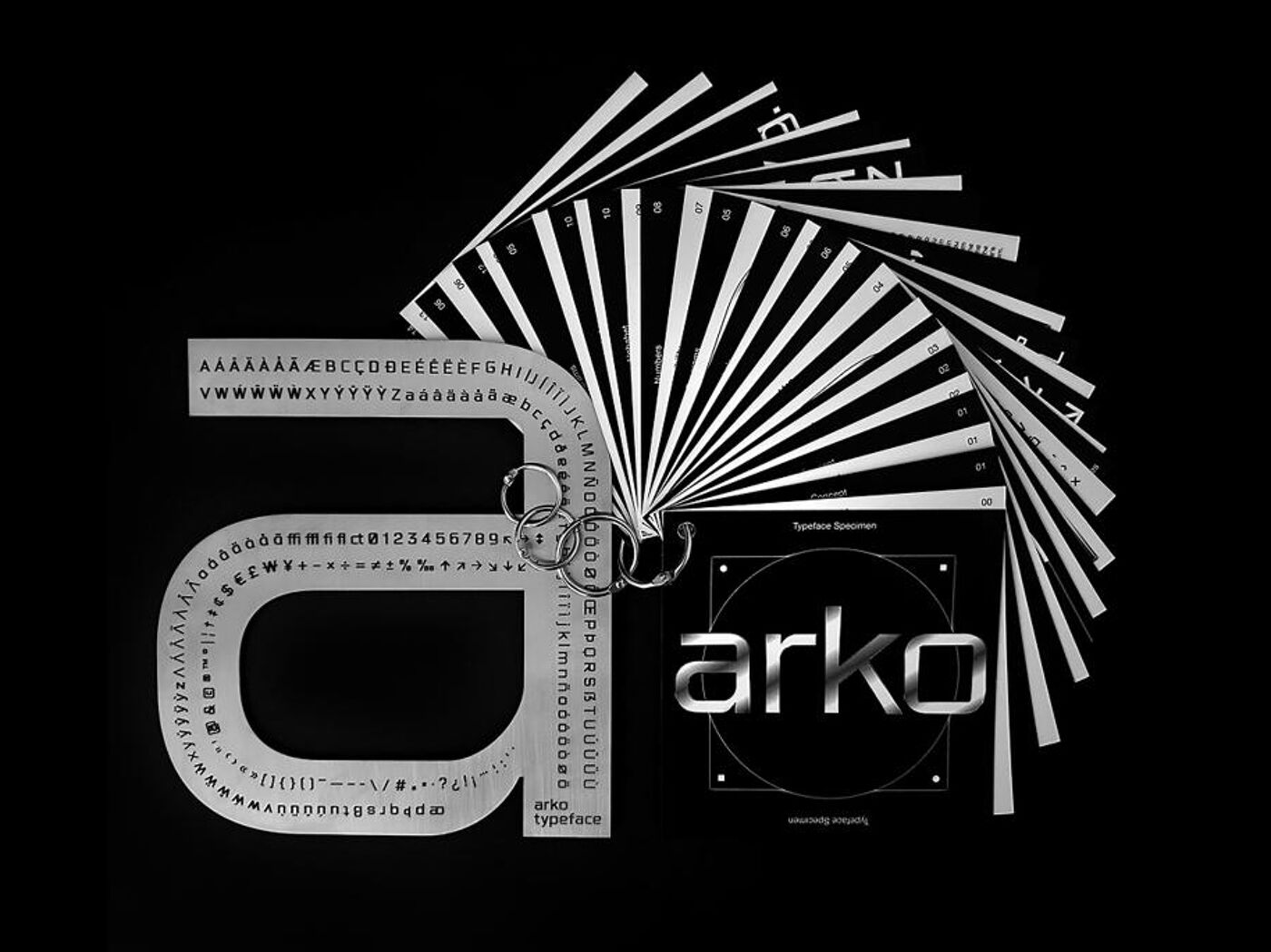 The image shows a black and white studio shot of Arko typeface design elements. A large stencil of the letter 'a' is prominently displayed next to the 'arko' logo, with several cards fanned out in the background, showcasing the typeface's various characters and symbols.