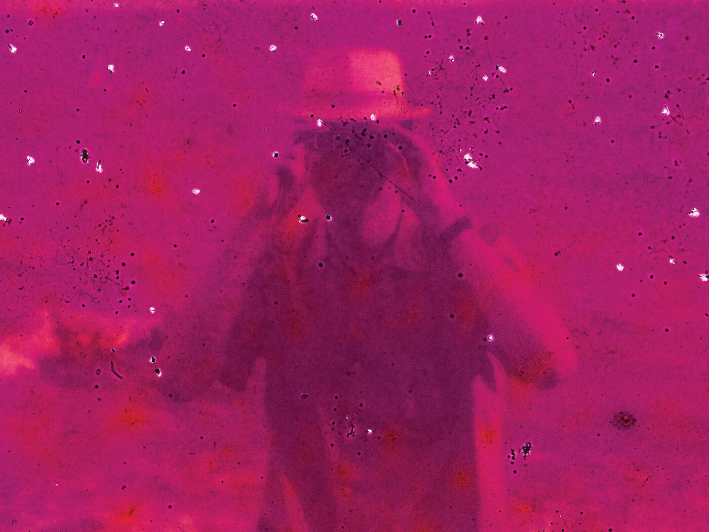 A person wearing a hat and sunglasses stands in an environment with a strong pink hue, possibly a desert or sandy area, with small white specks scattered around.