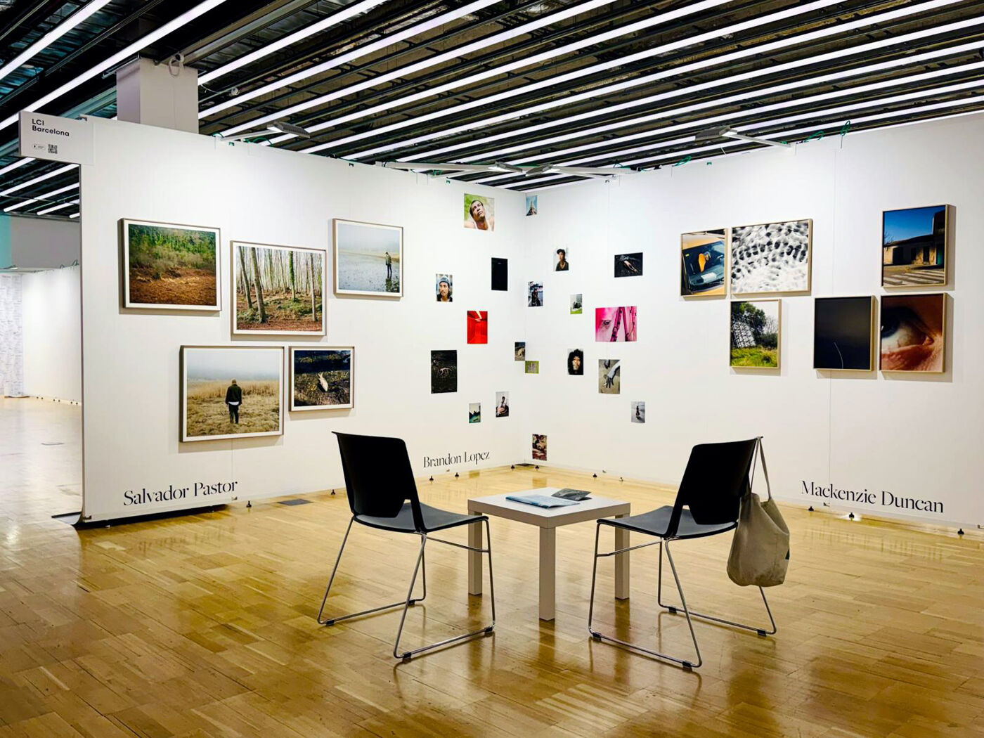 An art exhibition featuring various framed artworks on the walls, two black chairs, and a white table in the center, set against a polished wooden floor.