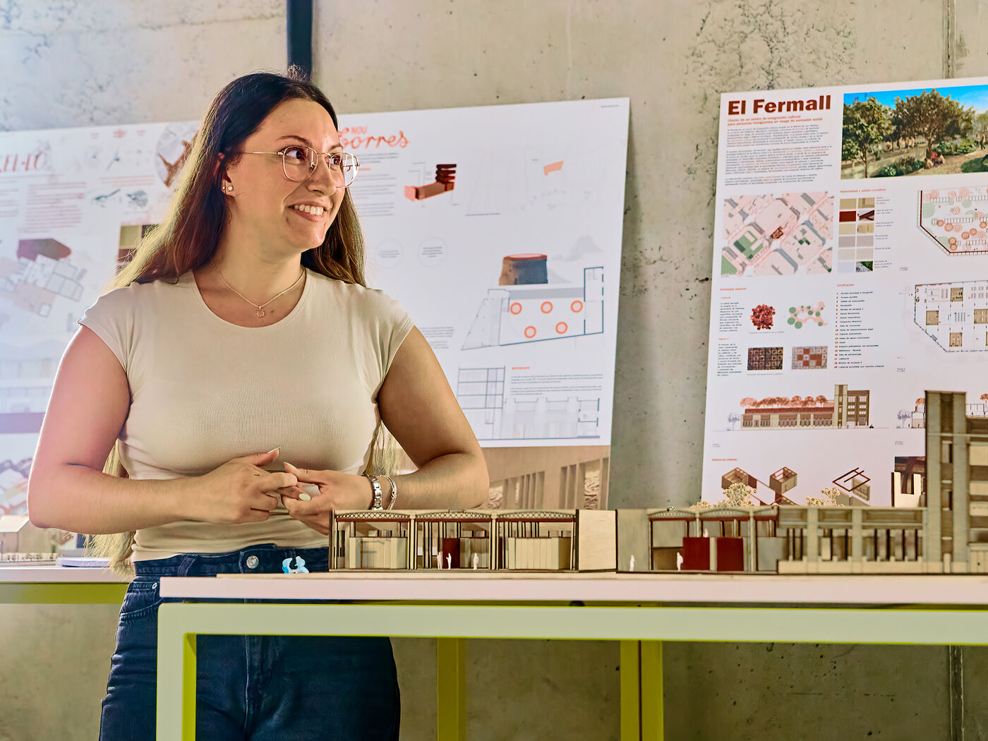 A smiling woman wearing glasses is presenting an architecture project with models and blueprints in the background.