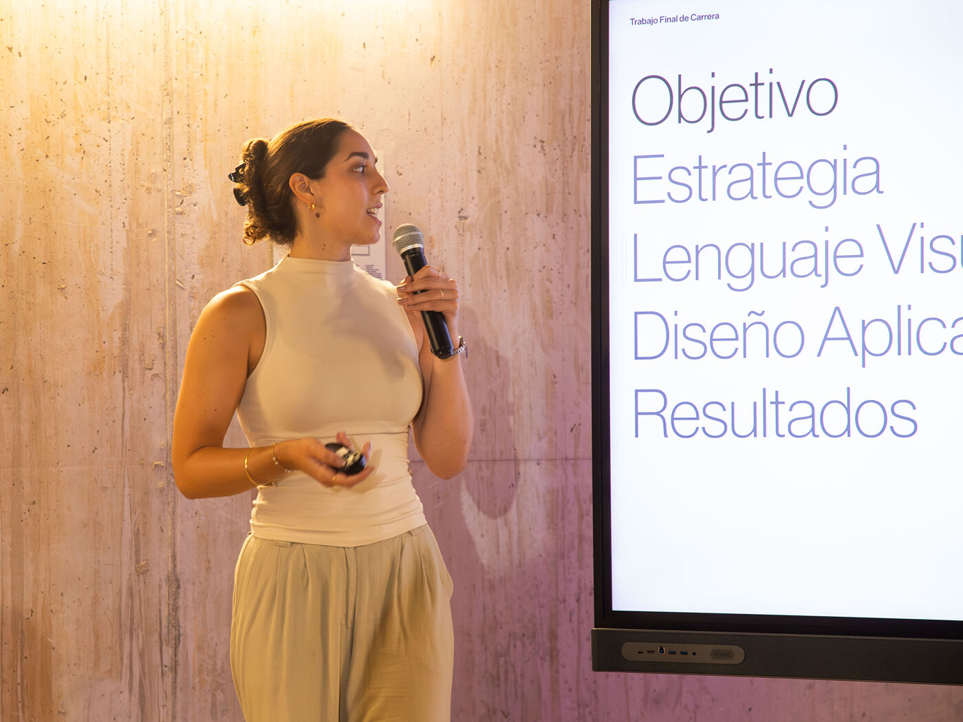 A woman is speaking at a conference, holding a microphone and a small device. To the right, a screen displays Spanish text including "Objective", "Strategy", "Visual Language", "Applied Design", and "Results".