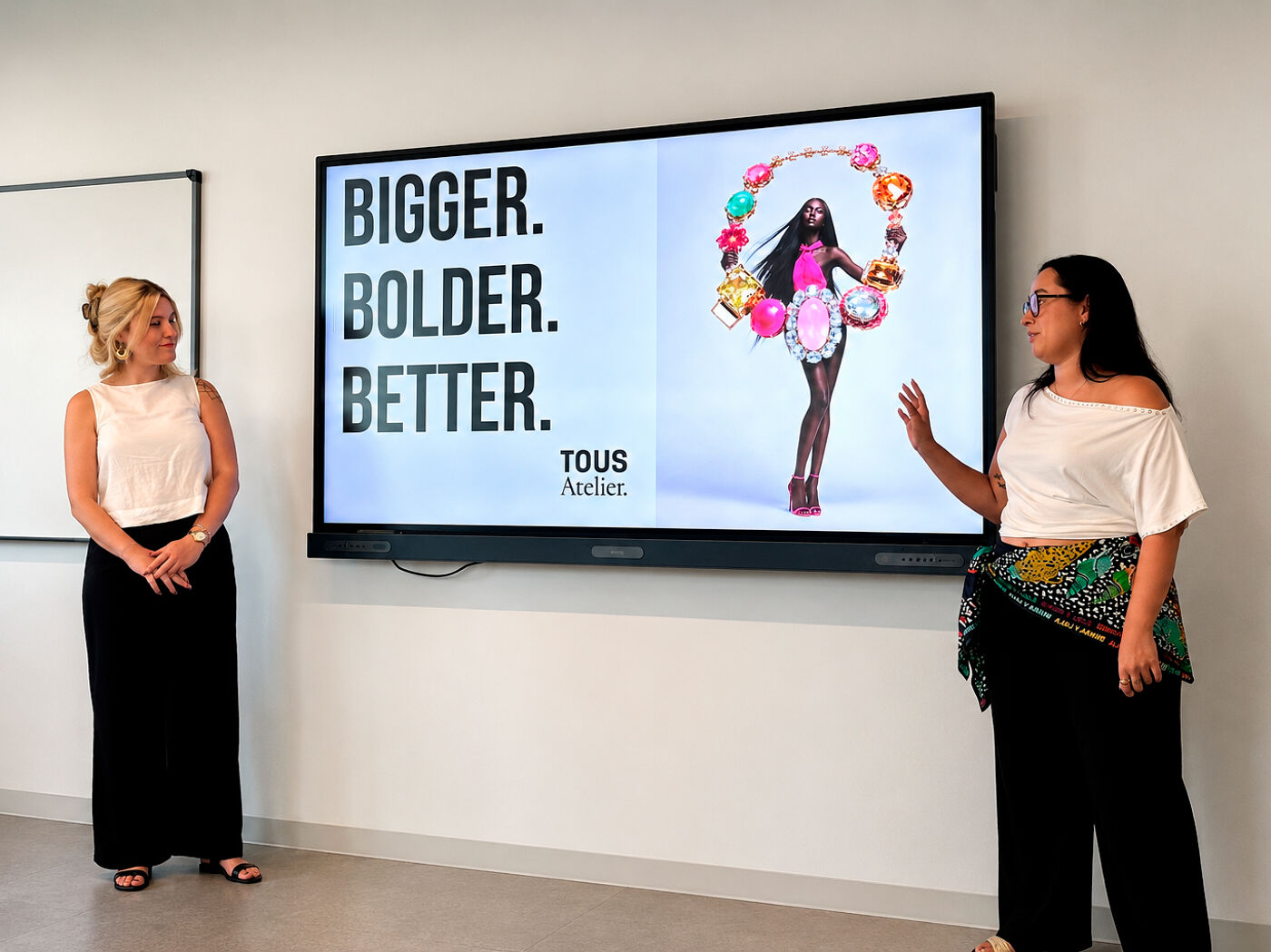 Two women stand before a large screen displaying "BIGGER. BOLDER. BETTER." and an image of a model adorned with vibrant jewelry and attire.