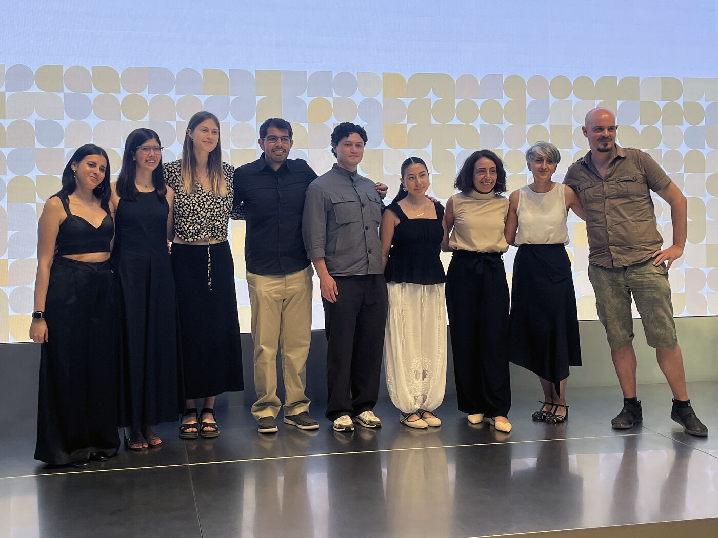 A group of nine people, both men and women, are standing on a stage in front of a backdrop with circular patterns. It appears to be an event or celebration.