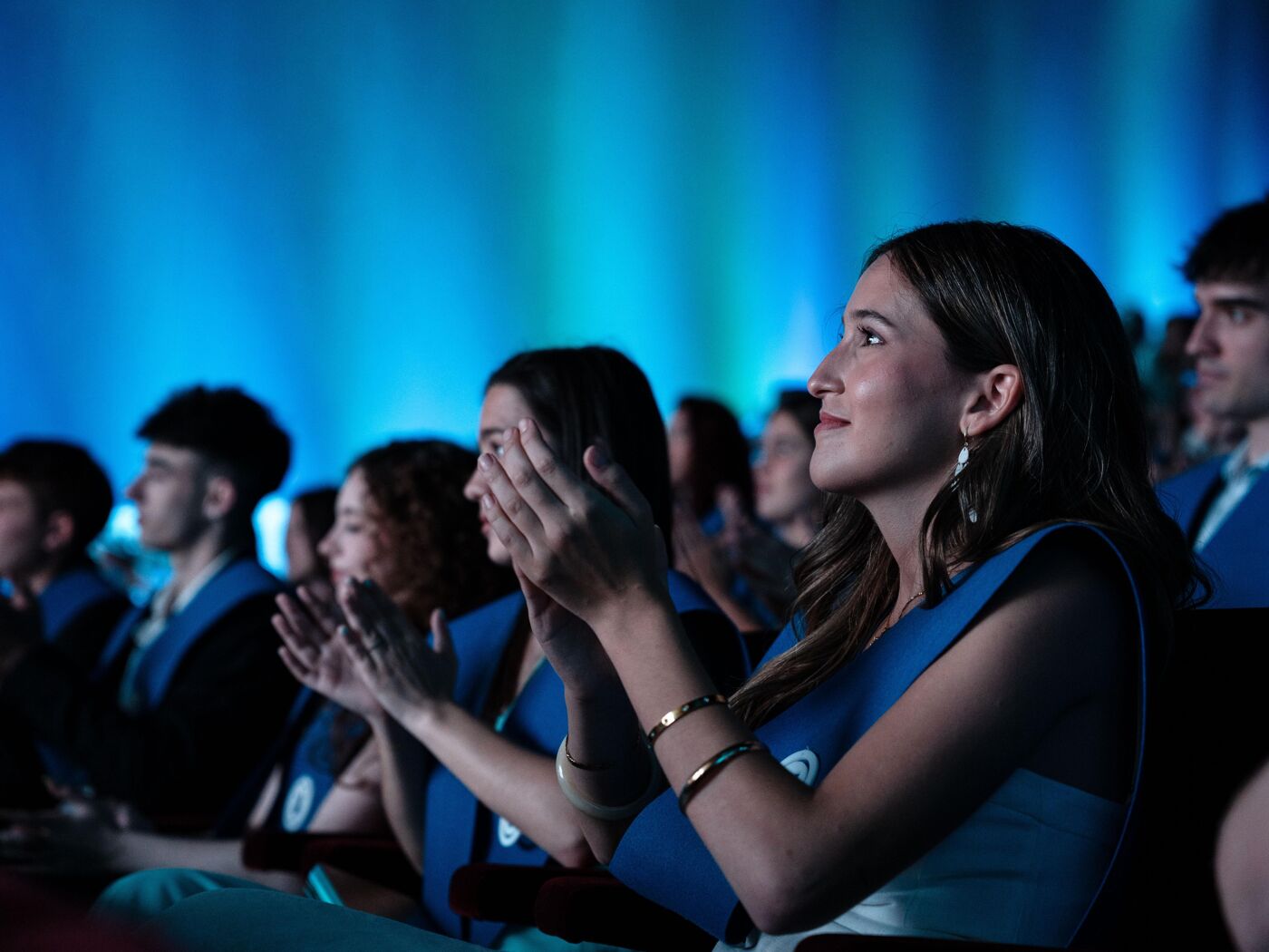 A group of people are clapping at an event, possibly a graduation ceremony. They are all wearing blue gowns with blue lights in the background.