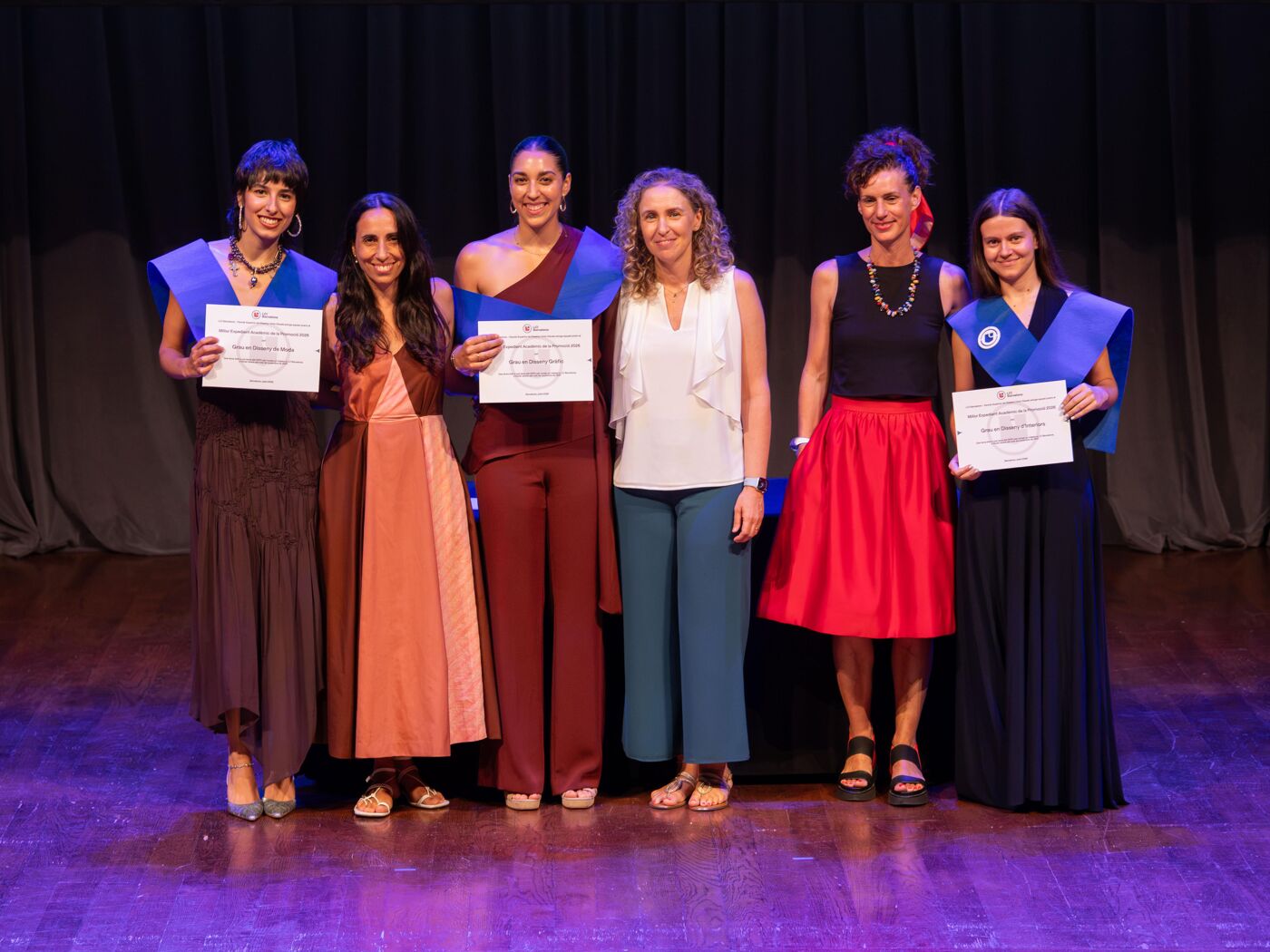 Six women pose for a photo on a stage, some holding certificates and others wearing blue and purple academic gowns.