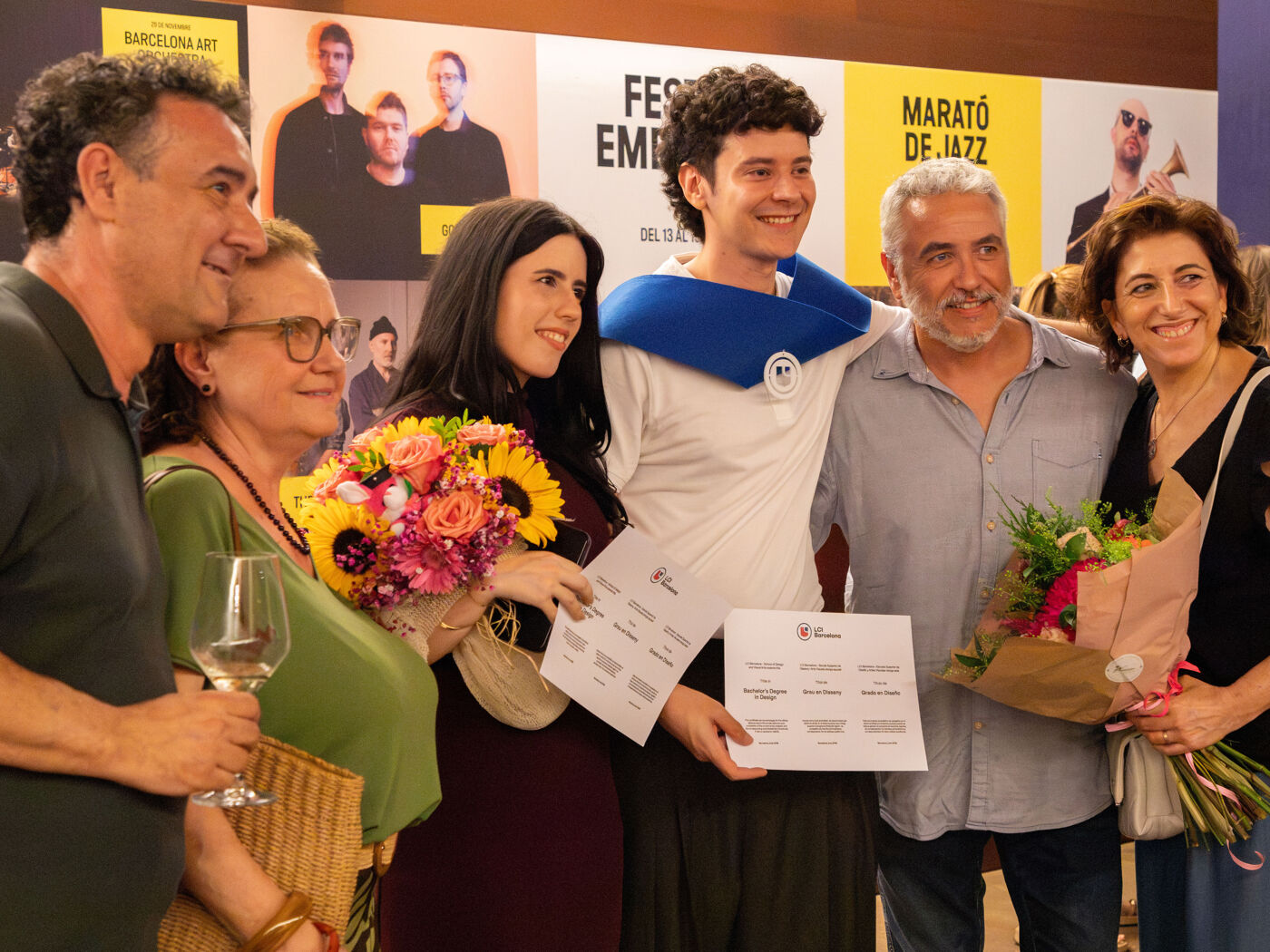 A group of people smile at the camera, some holding certificates and bouquets of flowers, celebrating an achievement at an event.
